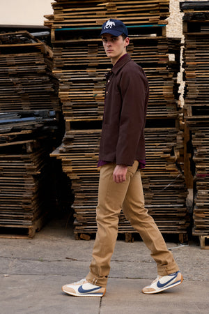A man walking in profile wearing a dark brown button-up shirt, tan pants, a navy baseball cap, and Nike sneakers, set against a background of stacked wooden pallets.