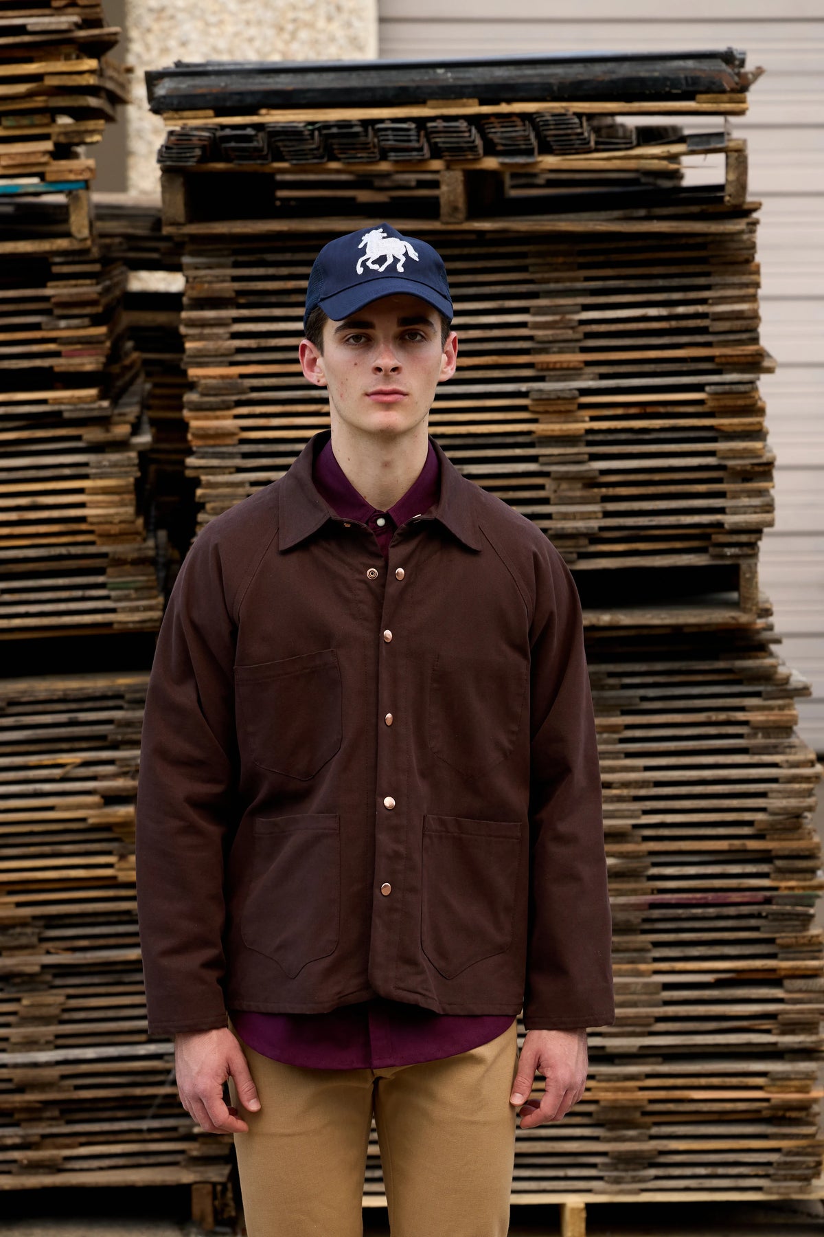 A man wearing a dark brown waxed cotton overshirt, a dark purple collared shirt underneath, tan pants, and a navy blue baseball cap with a white horse logo, standing in front of a stack of wooden pallets.