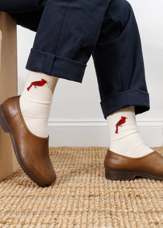 Person wearing white socks with red bird designs, brown shoes, and navy pants on a textured floor.