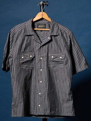 A short-sleeve button-up shirt with vertical dark gray and white stripes, hanging on a wooden hanger against a dark blue background. The shirt features two buttoned chest pockets and a camp-collar style neckline.