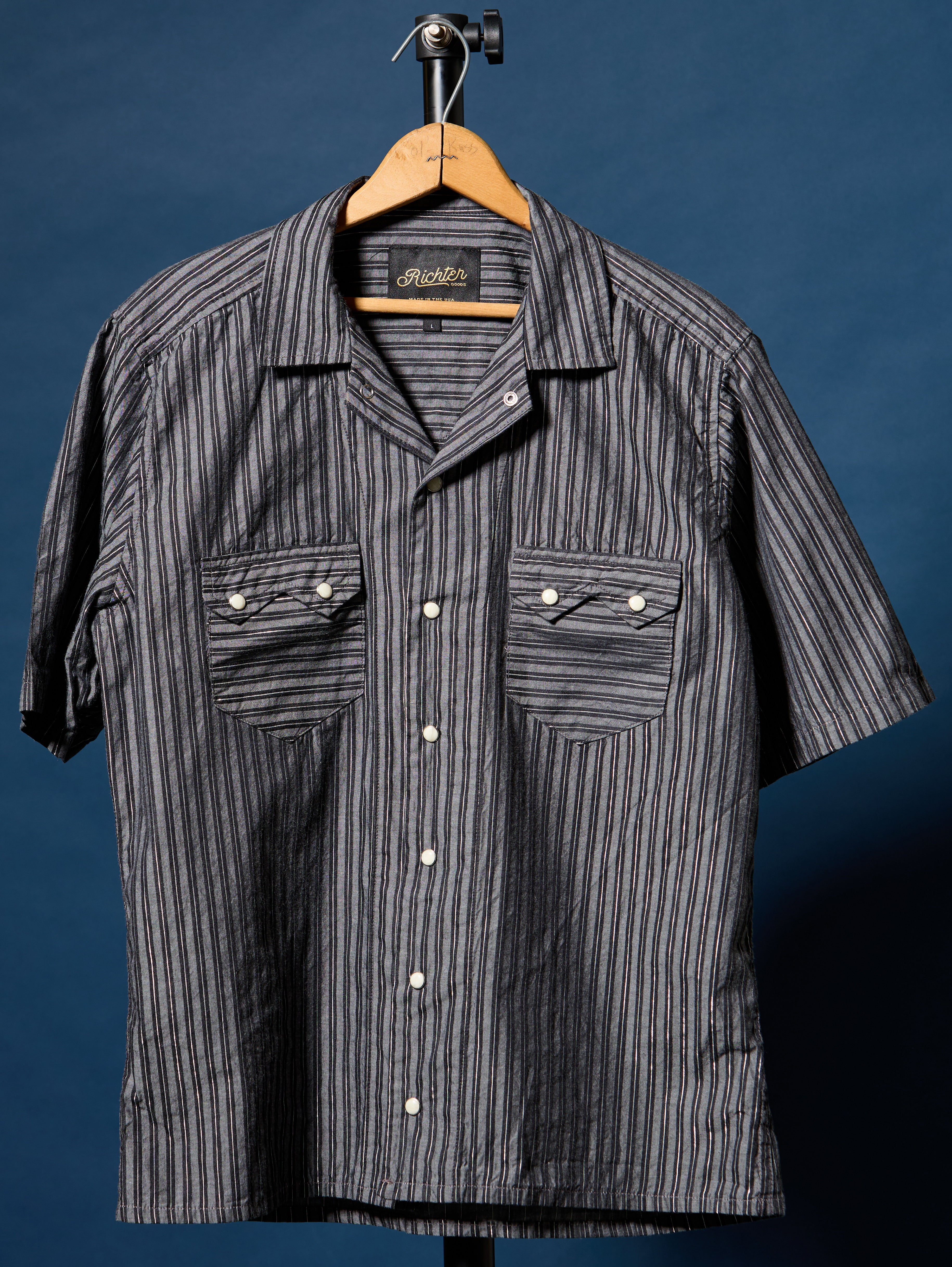 A short-sleeve button-up shirt with vertical dark gray and white stripes, hanging on a wooden hanger against a dark blue background. The shirt features two buttoned chest pockets and a camp-collar style neckline.