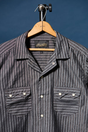 A gray and black vertically striped button-down shirt by Richter, hanging on a wooden hanger against a dark blue background.