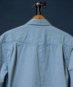 The back of a light blue linen button-up shirt hanging on a wooden hanger with a black clamp against a dark navy background.
