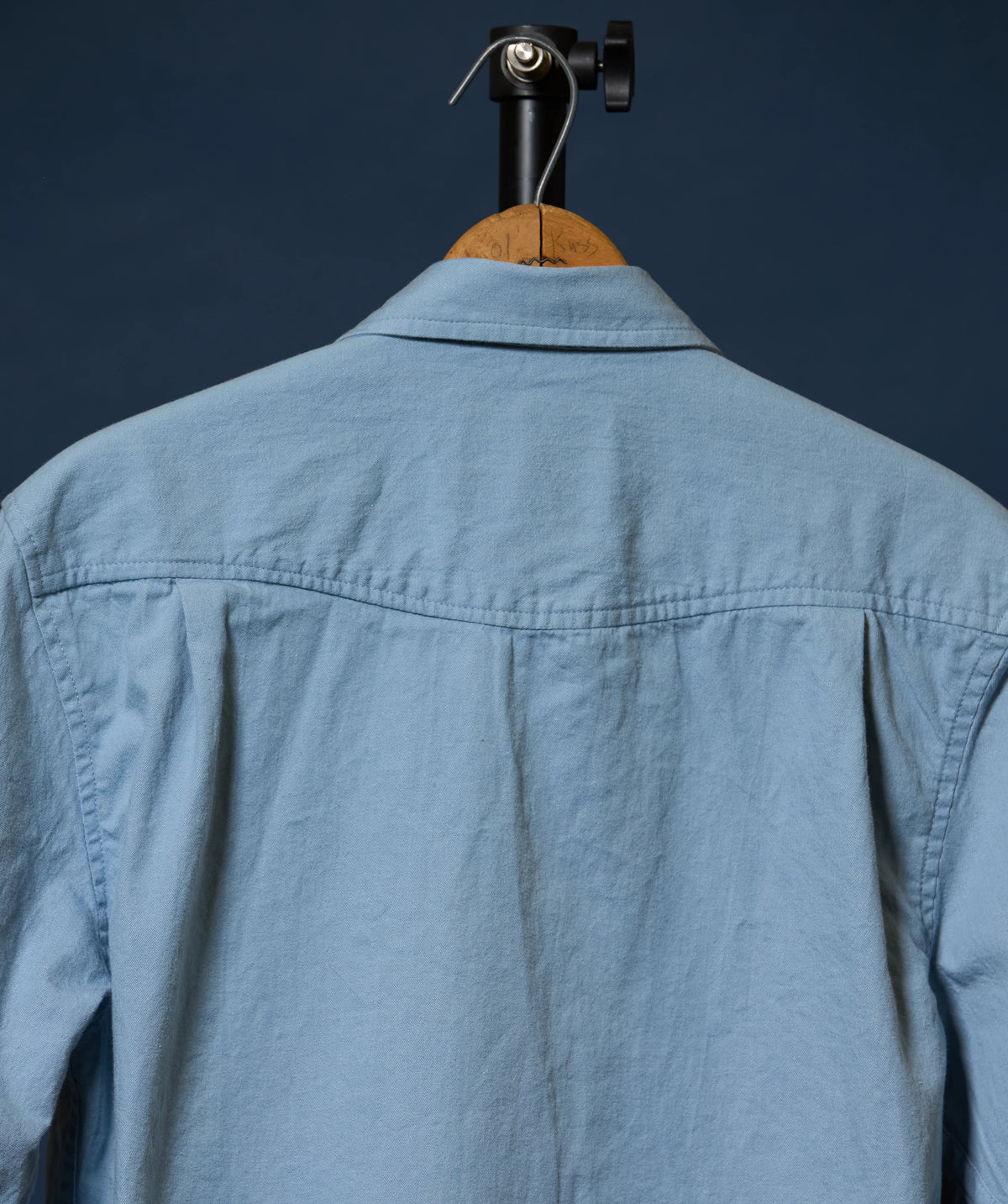 The back of a light blue linen button-up shirt hanging on a wooden hanger with a black clamp against a dark navy background.