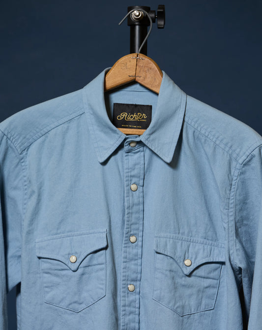 A light blue long-sleeve button-up shirt with two chest pockets, hanging on a wooden hanger against a dark navy background. The brand label inside the collar reads 'Richter'.