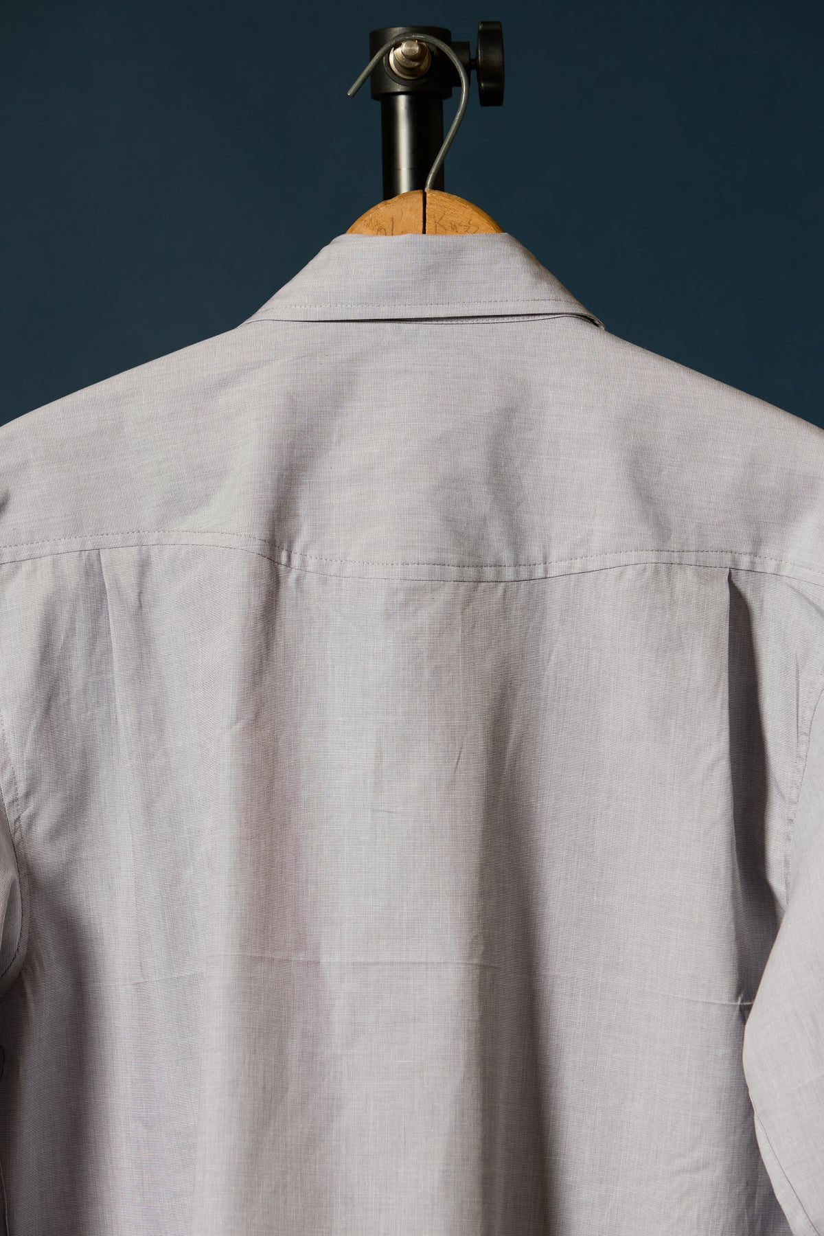 Close-up of the back of a beige long-sleeve button-up duck canvas shirt hanging on a wooden hanger against a dark blue background.
