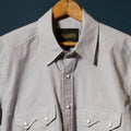 A Beige long-sleeve button-down shirt by Richter Goods, hanging on a wooden hanger against a dark navy background. The shirt features two buttoned chest pockets and a black brand label with gold lettering.
