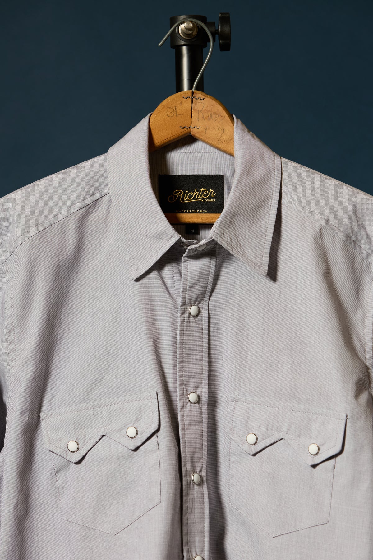A Beige long-sleeve button-down shirt by Richter Goods, hanging on a wooden hanger against a dark navy background. The shirt features two buttoned chest pockets and a black brand label with gold lettering.