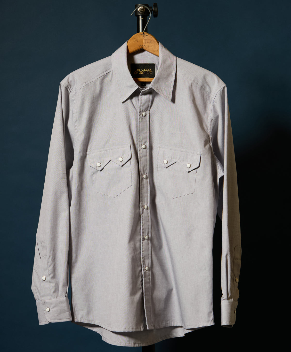 A Beige long-sleeve button-up shirt with two chest flap pockets, hanging on a wooden hanger against a dark blue background.
