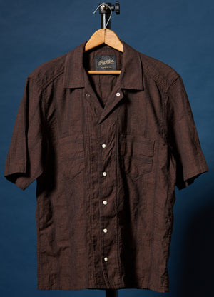 A dark brown short-sleeve button-up shirt with a textured crinkle fabric, hanging on a wooden hanger against a dark blue background.