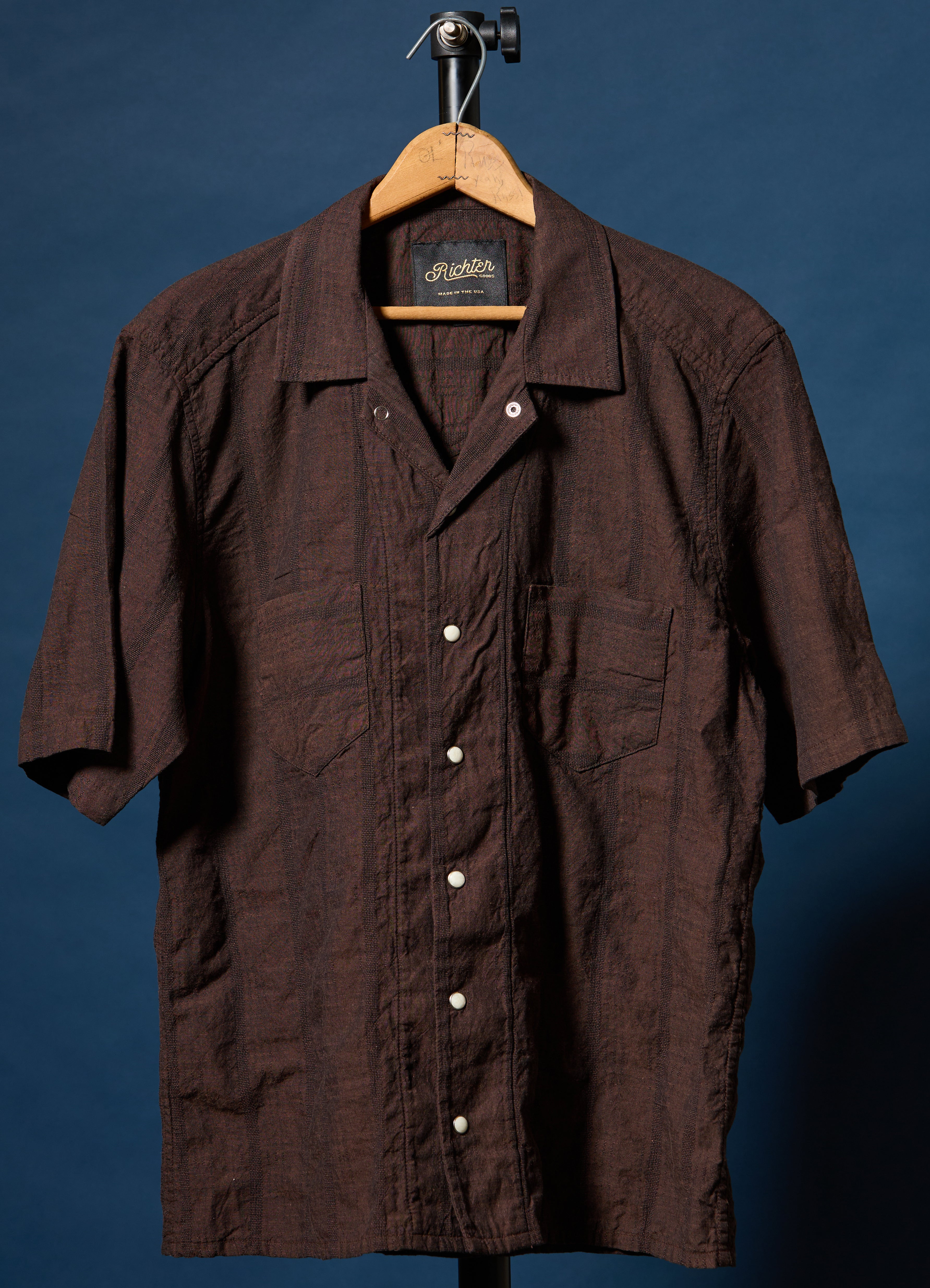 A dark brown short-sleeve button-up shirt with a textured crinkle fabric, hanging on a wooden hanger against a dark blue background.