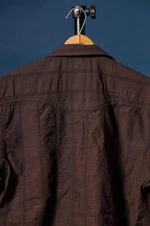 A dark brown textured cotton shirt hanging on a wooden hanger with a black metal hook against a solid dark blue background.