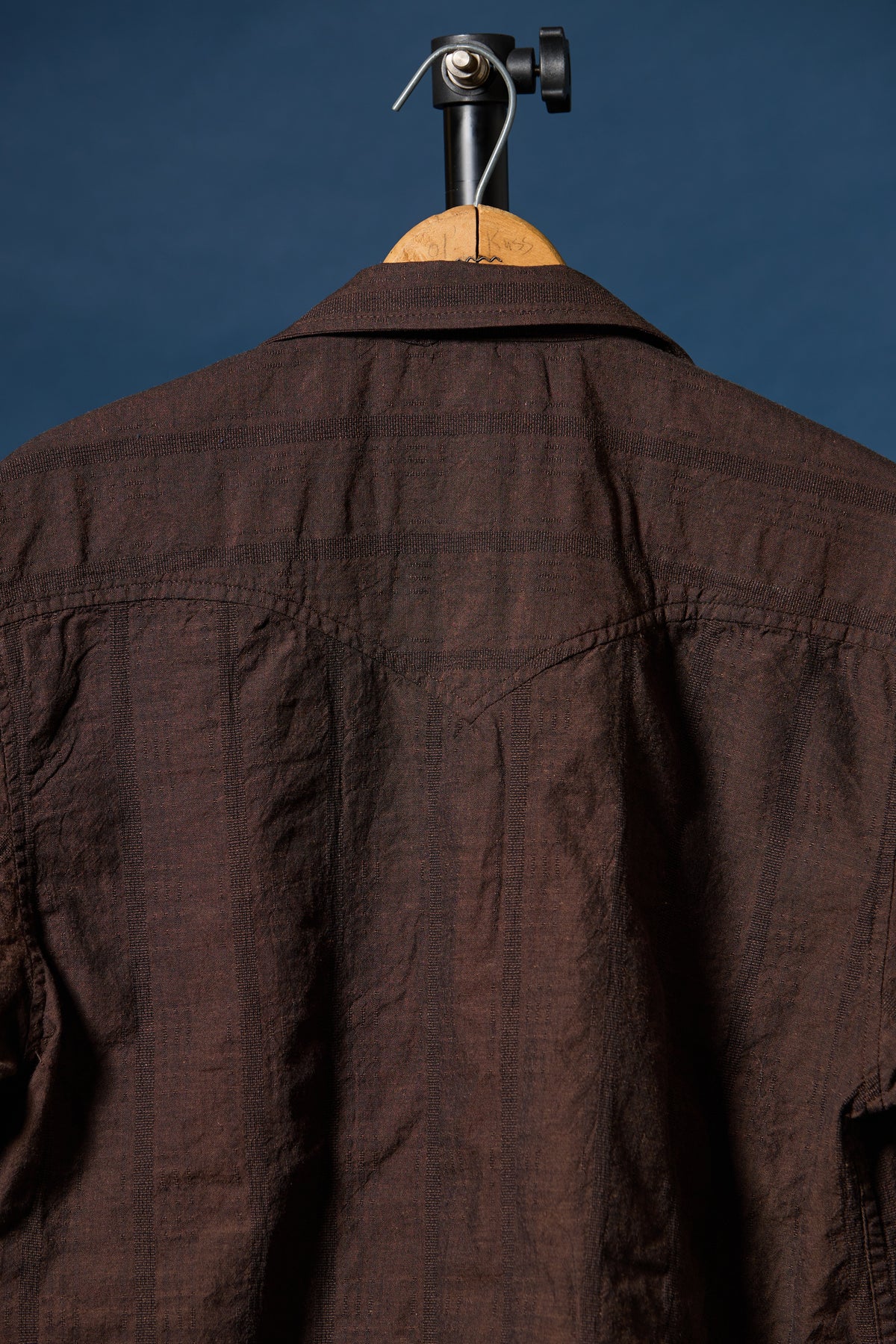 A dark brown textured cotton shirt hanging on a wooden hanger with a black metal hook against a solid dark blue background.
