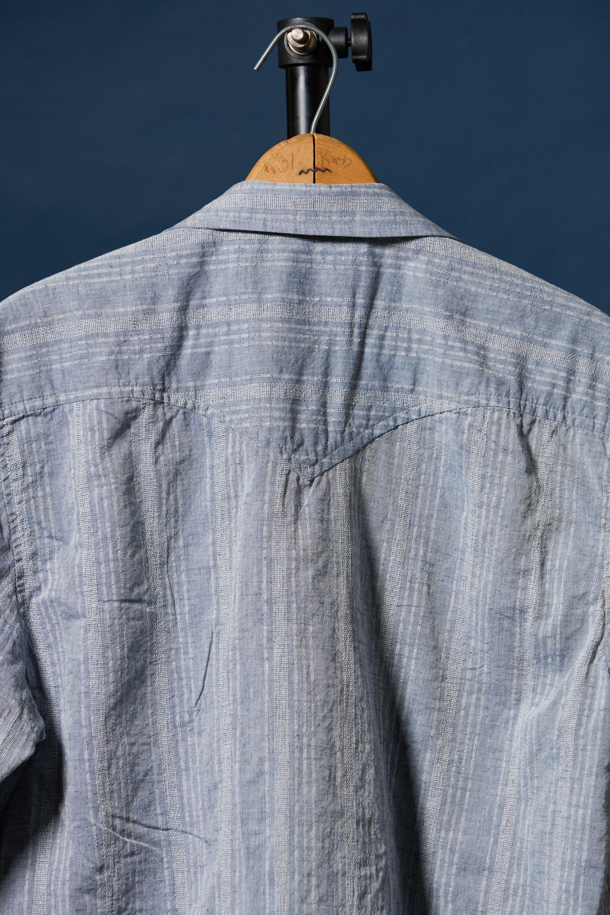 A light blue and white vertically striped cotton shirt hanging on a wooden hanger against a dark navy blue background.