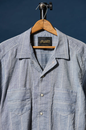 A light blue and white striped button-down shirt hanging on a wooden hanger against a dark blue background. The shirt features a camp collar, a button-down front, and two chest pockets.