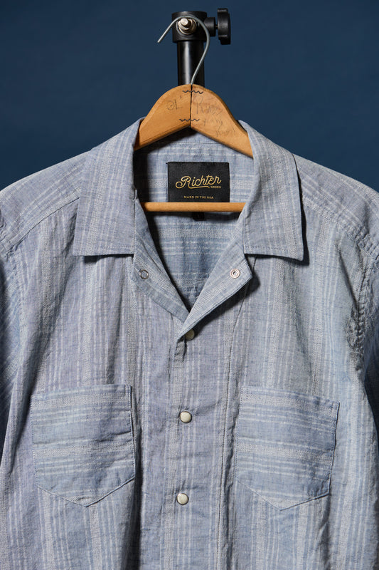 A light blue and white striped button-down shirt hanging on a wooden hanger against a dark blue background. The shirt features a camp collar, a button-down front, and two chest pockets.